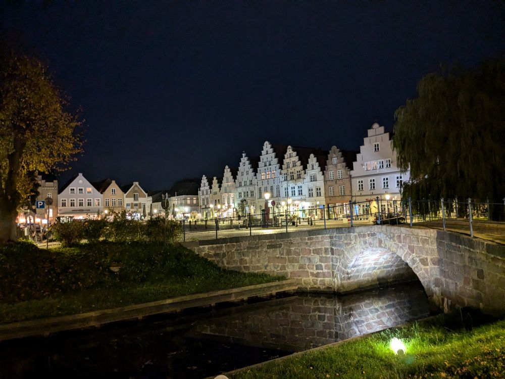 Zu sehen ist der Marktplatz von Friedrichstadt in Nordfriesland. Es ist sehr dunkel. Im Vordergrund sieht man eine kleine Steinbrücke die über eine Gracht führt. In der Mitte des des Bildes sind Häuser mit Stufengiebeln die durch Straßenlaternen beleuchtet werden. Der Himmel ist tief dunkelblau.