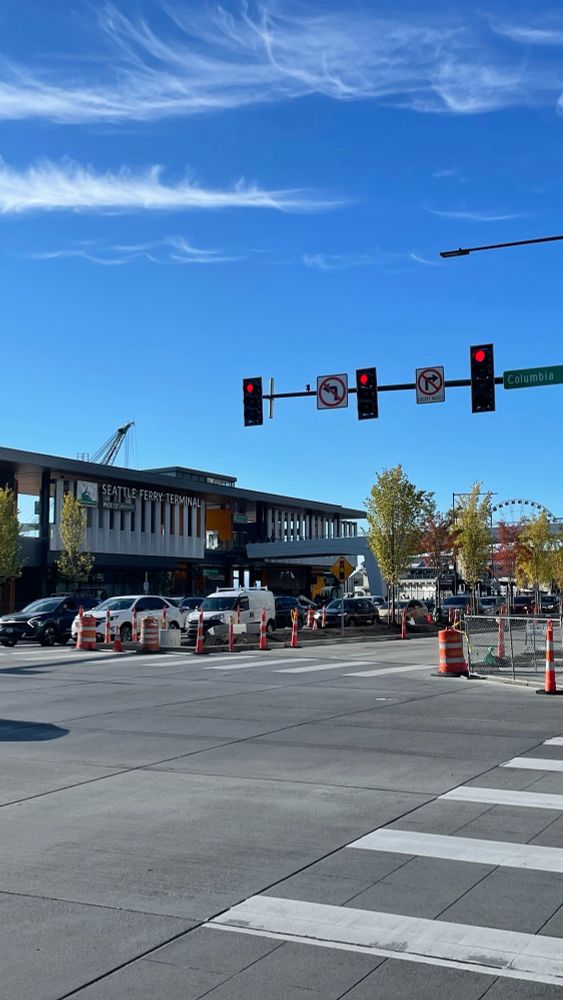 Das Bild zeigt eine Straßenszene mit einer Ampel im Vordergrund und dem Seattle Ferry Terminal im Hintergrund. Die Straße ist von Fahrzeugen, darunter Autos und Baufahrzeuge, sowie von einigen Fußgängern belebt. Im Himmel sind zarte, wellenförmige Wolken zu sehen, und es ist ein klarer, sonniger Tag.