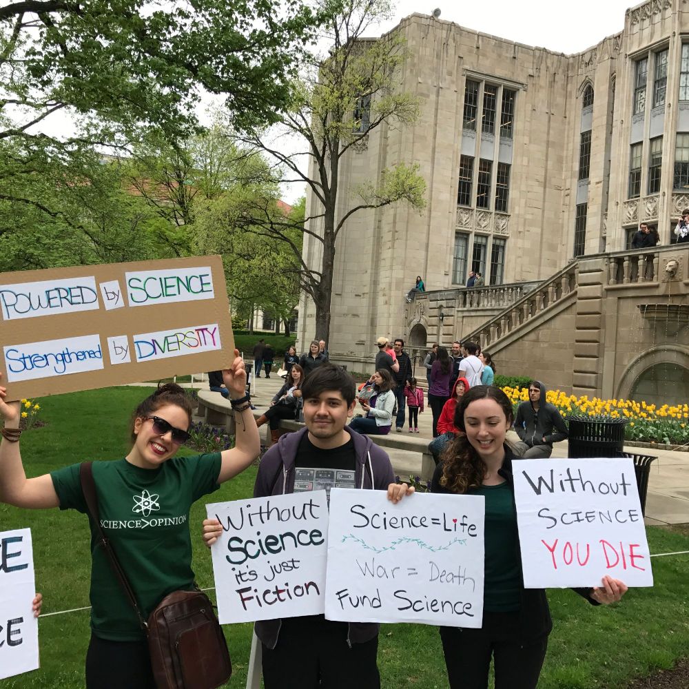 a photo of scientists with protest signs that read: 1) Powered by science strengthened by diversity, 2) without science its just fiction, 3) science = life war = death; fund science 4) without science you die