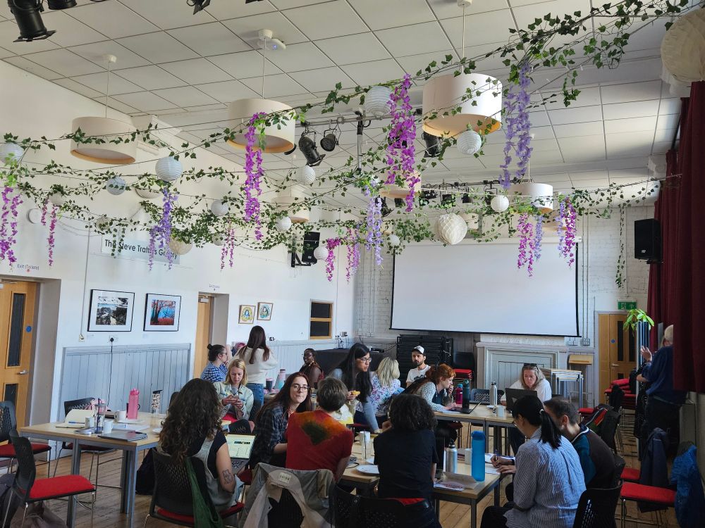 A large meeting room inside, woth lots of members of the Centre for Law and Social Justice across various tables. Floral green and lilac buying cross crosses pleasantly overhead 