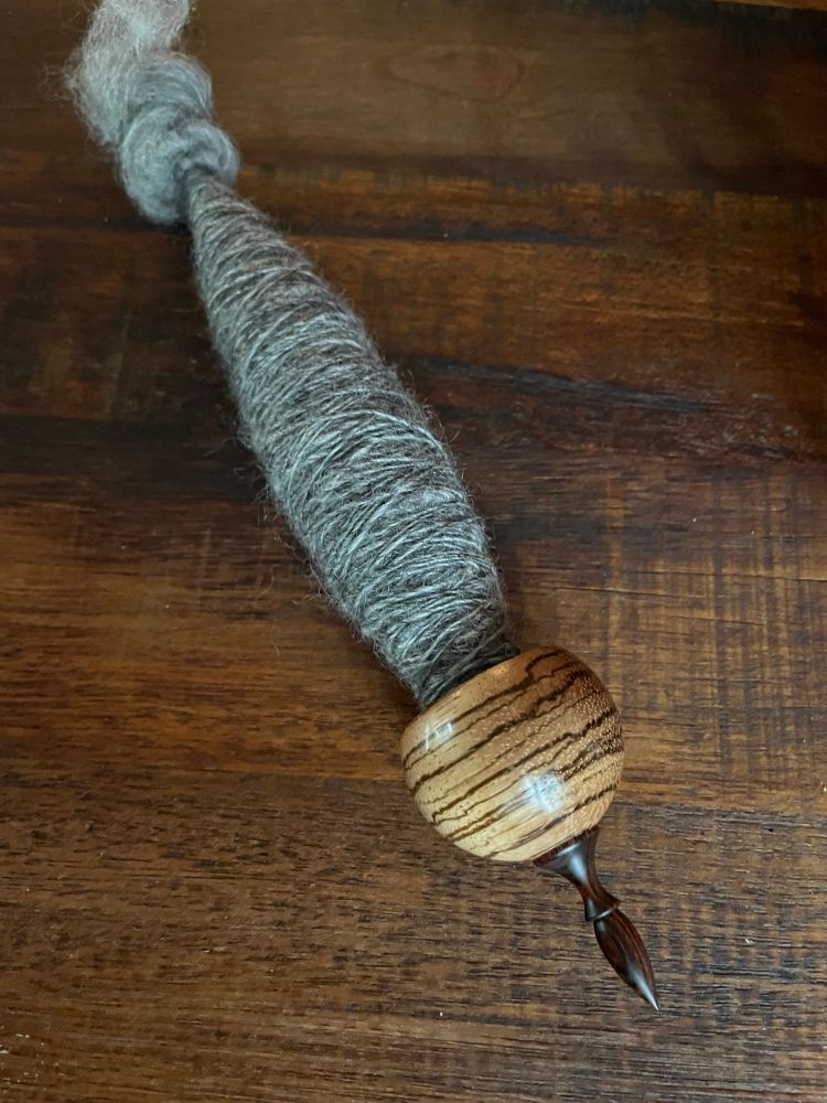 A zebra wood support spindle on a rustic wood background. The spindle has a single of gray textured fiber wrapped around the shaft.