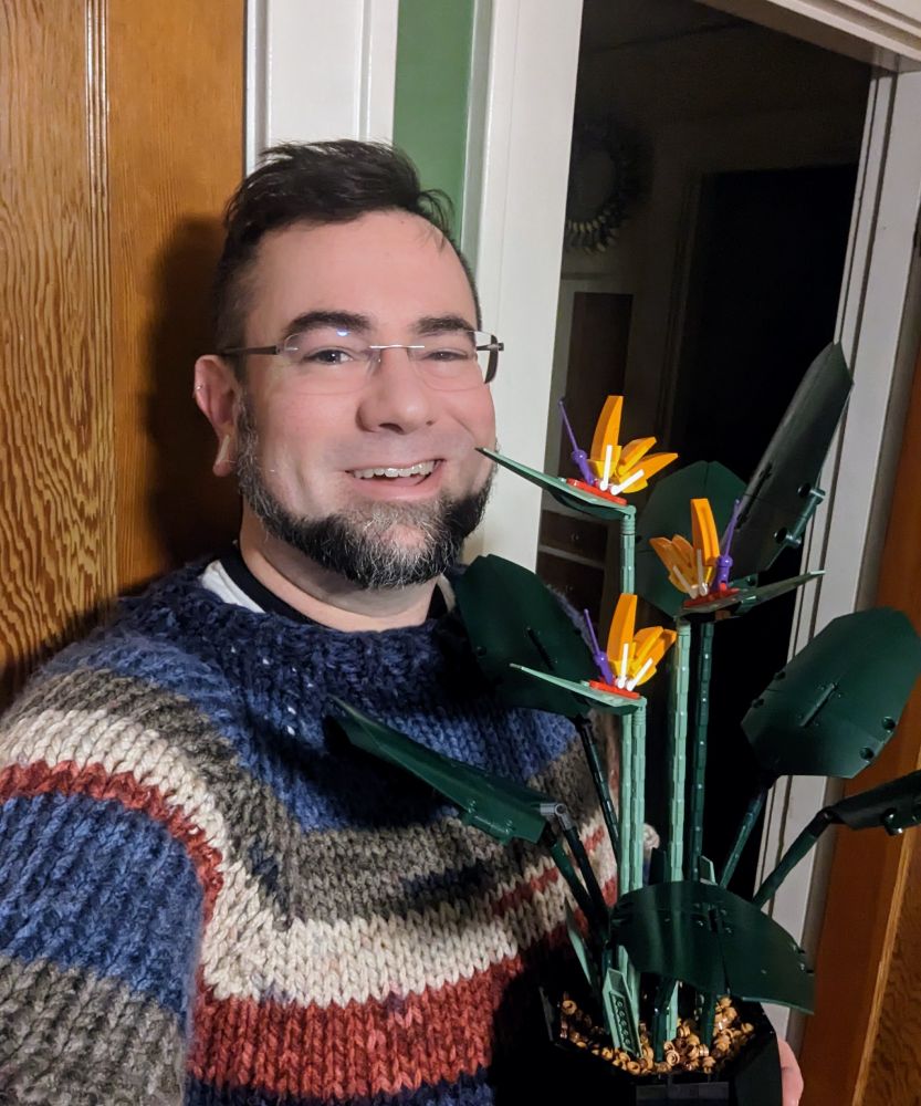 Steve, a middle-aged Caucasian man in glasses and a handmade sweater, smiles at the camera while holding a flower made of lego