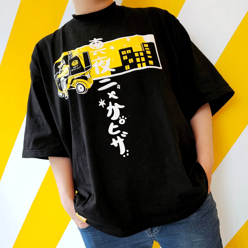 An oversized black Tshirt with yellow and white print. The print is a picture of a cat driving a pizza delivery bike in front of a night sky