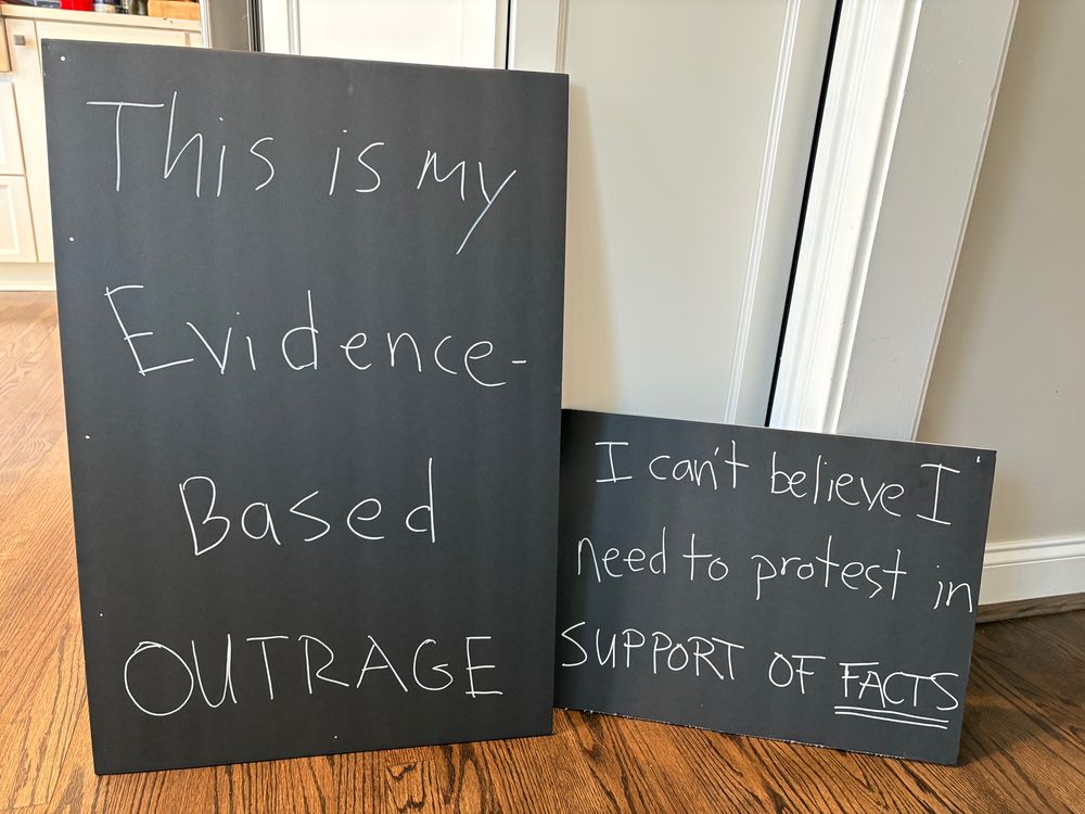 Two chalkboard-like signs saying “this is my evidence-based outrage” and “I can’t believe I need to protest in support of facts.”