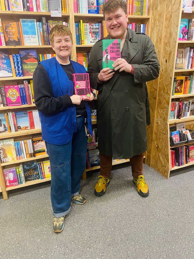 elizabeth and the author Jake Hall pose together with a copy of their books: THANK YOU FOR CALLING THE LESBIAN LINE and Shoulder to Shoulder in front of a bookshelf 