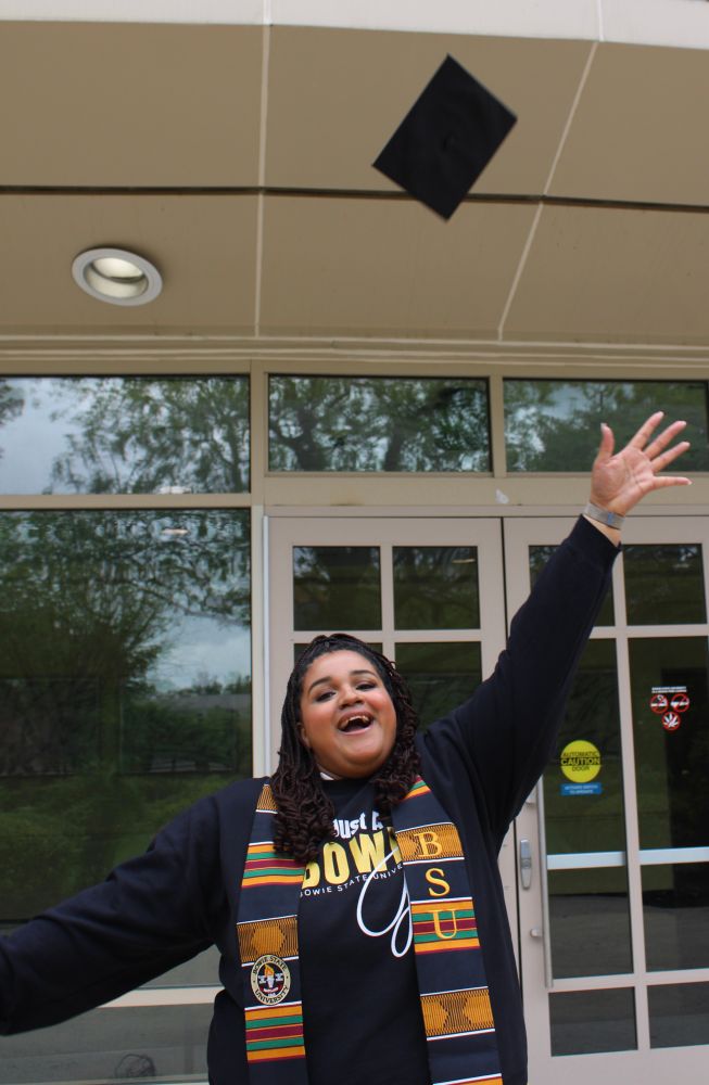 A photo of myself looking at the camera after I threw my grad cap in the air. The cap is in the air on the top right above my hand. I have both arms up in the air. I am wearing a Black sweatshirt with white and yellow lettering that says “just a Bowie Girl”, though some of it is covered by my hair and my stole. I am also wearing my HBCU kente stole that has the Bowie State logo on one side and BSU on the other side.