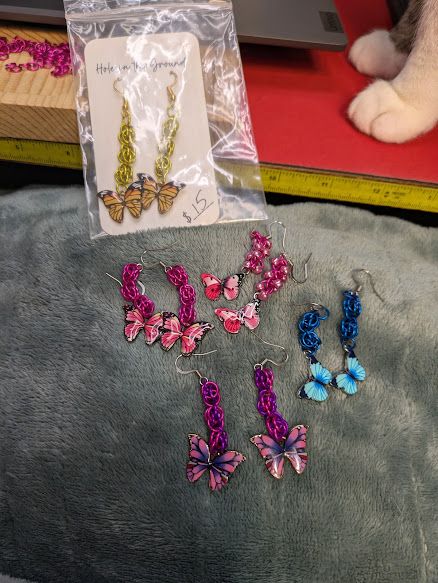 5 pairs of earrings. They each have butterflies as pendants, with chainmaille sections to make them dangly. 3 pair are pink. One pair is yellow. One pair is blue. There is also a pair of white kitty paws showing on the table behind them.