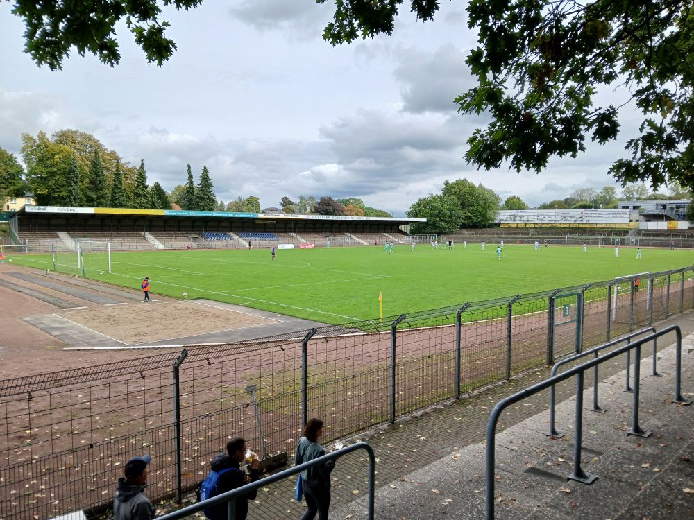 Abgebildet ist die Haupttribüne und Teile der Südkurve im Röntgen-Stadion in Remscheid, sowie das Spielfeld und Teile des Zauns auf der Gegengerade. Hinter der Haupttribüne stehen einige Bäume. 
