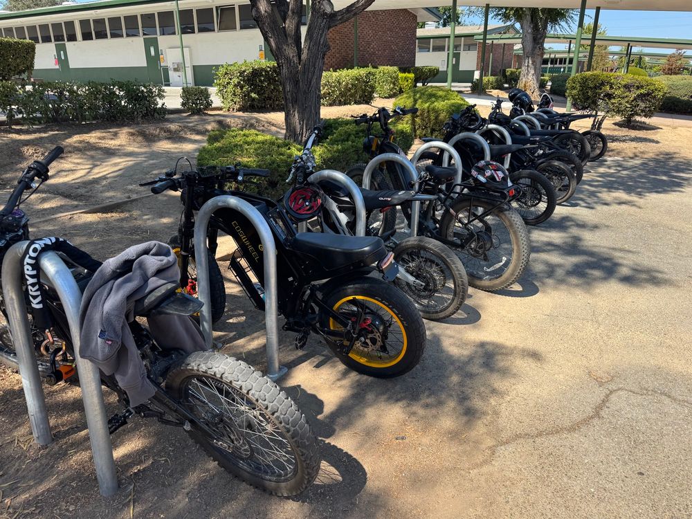 Photo of a row of bike locking racks on a school campus, filled with about a dozen heavy e-bikes or e-motos and no apparent “analog” bikes. 