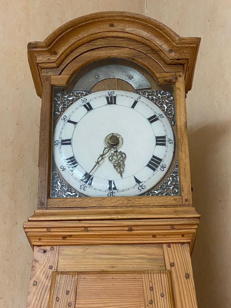 Zu sehen ist eine alte Standuhr aus dem Jahr 1793, die Zeiger stehen auf fünf Minuten nach halb sechs. Ein rundes, weißes Zifferblatt mit schwarzen römischen Zahlen, in den Ecken des runden Zifferblatt zum eckigen Gehäuse sind silbrige Verzierungen. Der Korpus aus Holz ist noch original, eine im 19. Jahrhundert aufgetragene bräunliche Farbe wurde in den 1980er Jahren abgebeizt. Die Uhr funktioniert immer noch einwandfrei mit Hilfe von Gewichten aus Blei zum Aufziehen. 