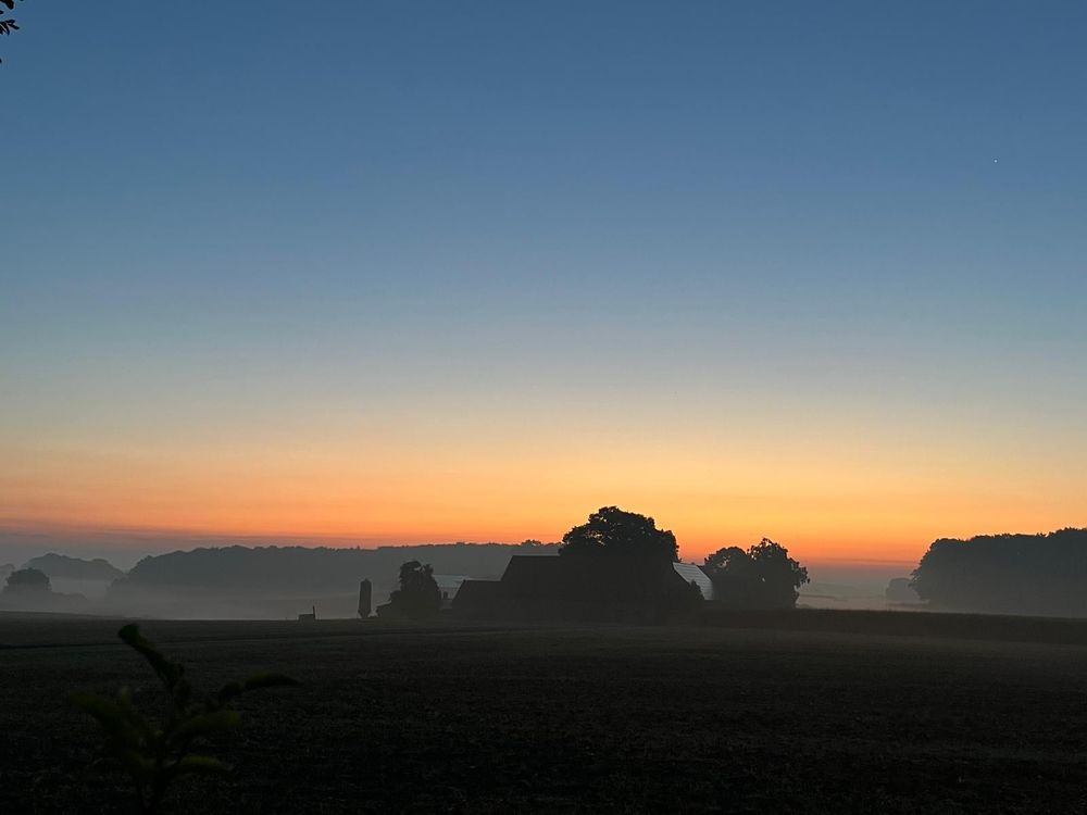 Sonnenaufgang im südlichen Osnabrücker Land. Bodennebel liegt auf dem Land, mittig ein Gehöft umgeben von Bäumen. Der Horizont mit den ersten orangefarbenen Vorboten der Septembersonne. 