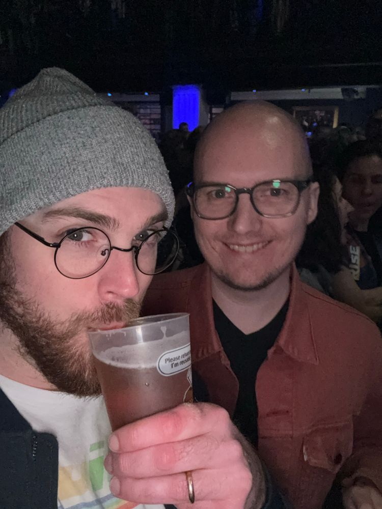 I sip on a beer on the left, my husband smiles on the right. We are in a dark crowded venue, anticipating the start of Ani Difranco’s performance. On the left my upper body is visible, I have a sparse beard, circular glasses, and a grey hat. On the right, my husband smiles in his rusty orange jacket and a black t shirt. He wears glasses as well, but his shaved head is uncovered. We are mildly surprised by the flash of the camera.