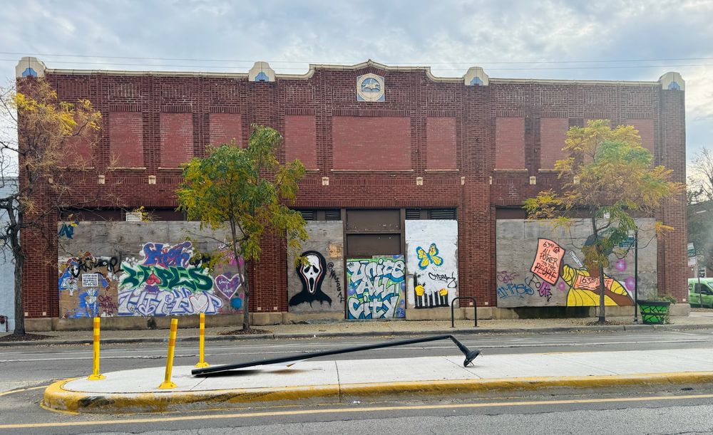 Color photo of a the front elevation of a 2 story red brick building taken from across the street. The first floor has been boarded up, but street artists have enlivened these makeshift canvases with murals. Above the second floor, the blue and white circular postal seal is centered, with additional modest blue and white terra cotta ornament spaced evenly across the roofline. 