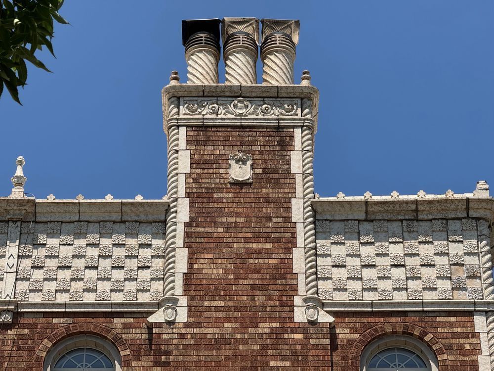 Color detail of ornament surrounding an ornate chimney of variegated brown brick. The chimney is ornamented by twisting columns and quoins on either side, and a small frieze at top featuring some type of foliate designs. On top of THIS are three chonky twisty chimney pots. On either side of the chimney continues the incredible band of terra cotta that is made of alternating plain tiles and raised tiles with rosettes in a checkerboard pattern. 