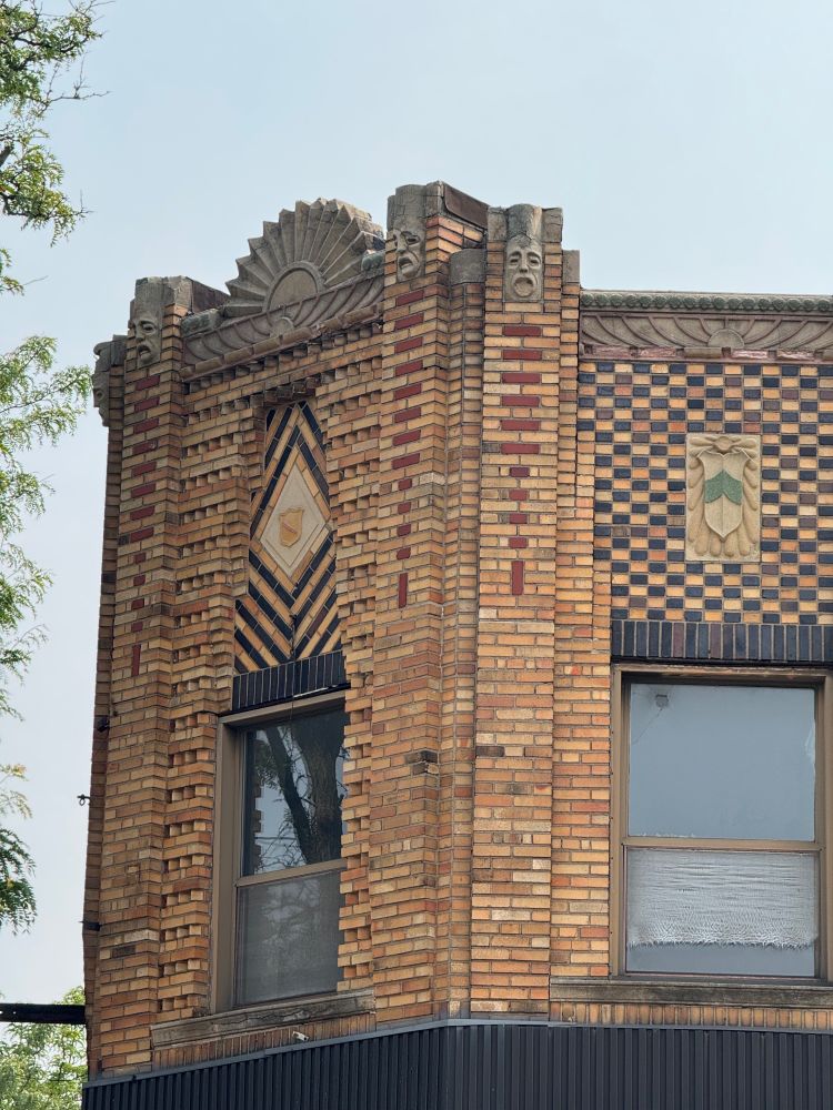 Color close-up of the second floor above the former corner entrance, and a small portion of one of the side elevations. On the corner, a concrete sunburst sits above the the parapet, with faces staring out from atop piers that are themselves ornamented by a red brick pattern giving this two story building a sense of verticality. Underneath the sunburst is ANOTHER sunburst, this one a more stylized Art Deco-y version. Centered underneath this is a panel with a golden shield bracketed by an arrangement of dark and light brick creating a diamond pattern highlighting the shield. On either side of this panel are alternating courses of stretchers with sawtooth brick, giving this tiny little section an amazing amount of visual interest and texture.