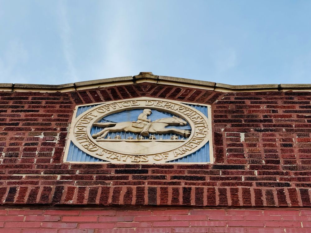 Color close-up of the USPS postal seal on a red brick building. The seal itself, featuring a well defined rider atop a sprinting horse on a blue background, is rendered in terra cotta. 