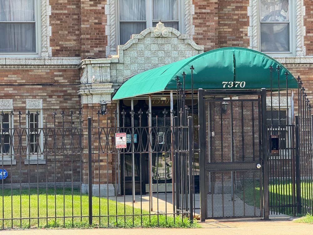 Color close-up of the entrance to the building, which features a large terra cotta pediment ornamented by scrolls and the same tile design seen along the top of the building. Unfortunately, much of this is obscured by a long green canopy that stretches from the door to the sidewalk, offering protection from both the elements and photography. Elsewhere in the photo can be observed ornate lintels and twisting columns along with quoins around the second floor windows.