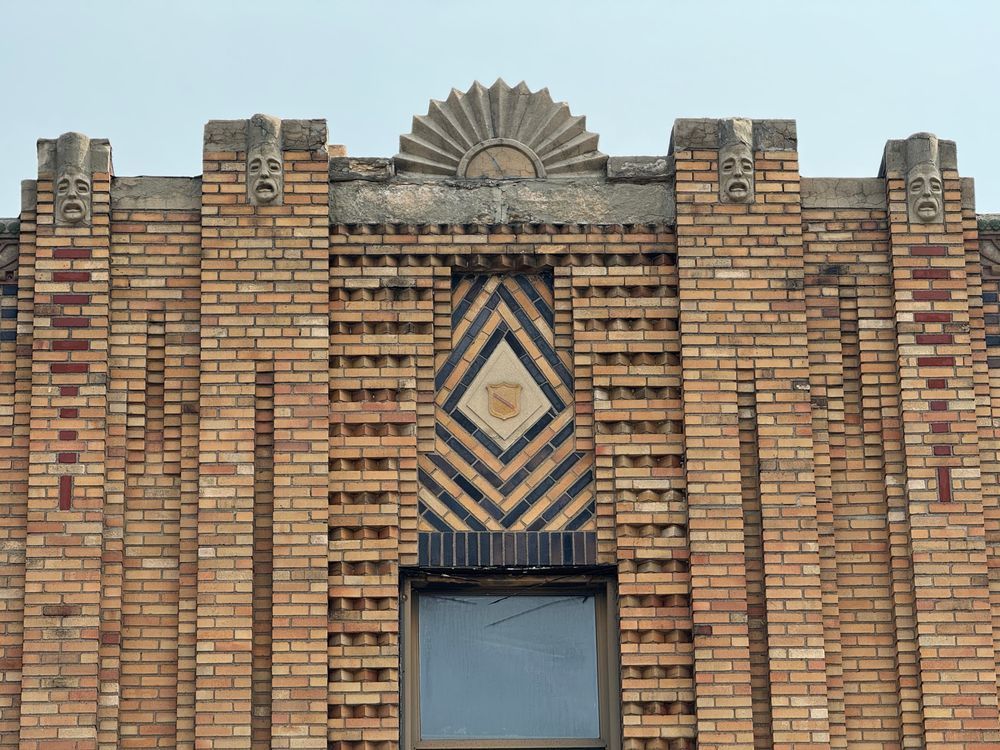 Color close-up of the brickwork around one of the windows on the side of the building. There's a lot going here. A cast stone sunburst! A shield set into a diamond brick pattern made from alternating dark and tan bricks! Sawtooth brick! Tan brick laid stretcher to stretcher! Red brick where they've used damn near every side of the brick (stretcher, header, soldier) to create a vertical pattern that serves as a visual arrow! Recessed brick arranged vertically! Faces!