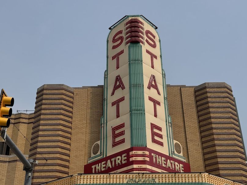 Color close-up of the giant, triangular vertical sign sitting above the marquee, taken from dead center in front of it. On either side, florescent red lights spell out STATE vertically above a red band with cream letters that read THEATRE. In tiny letters, along the top of the marquee are the words NOW PLAYING