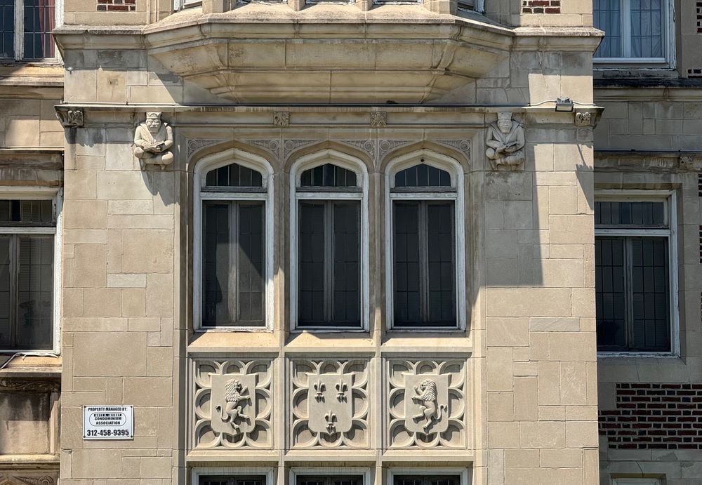 Color photo of a light stone building showing three windows, each with an arched transom, making them look lancet-ish. Above the windows can be seen the bottom of a bay, and underneath are Gothic spandrels featuring a shield sitting on a bed of blind tracery. The shields on the two outer spandrels have those bipedal lions that look like they're about to box (lion rampart), and the middle one has three fleurs-de-lis on it. At the top corner of each window is a carved little guy sitting crisscross applesauce since 1928 and from his expression, he's pretty unhappy about it.