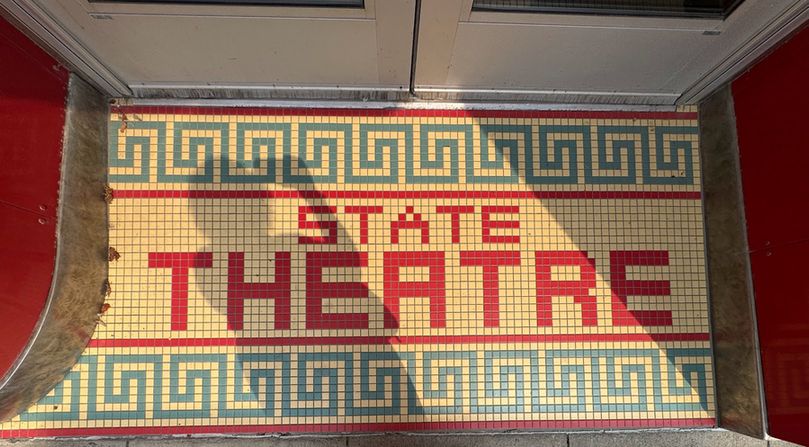 Color photo of 1-inch square tiles adorning one of the theater's entrances. On a background of cream tiles is the word STATE on top of the word THEATRE, both in red tiles. The word 'state' is spelled out using just one row of tiles, but the word 'theatre' uses two and so the word is consequently much larger. A line of red tiles separates these word from a light teal meander at both top and bottom. If this was in black and white, my shadow would fall film noir-ishly across half the tiles, but since the photo is in color please ignore it.
