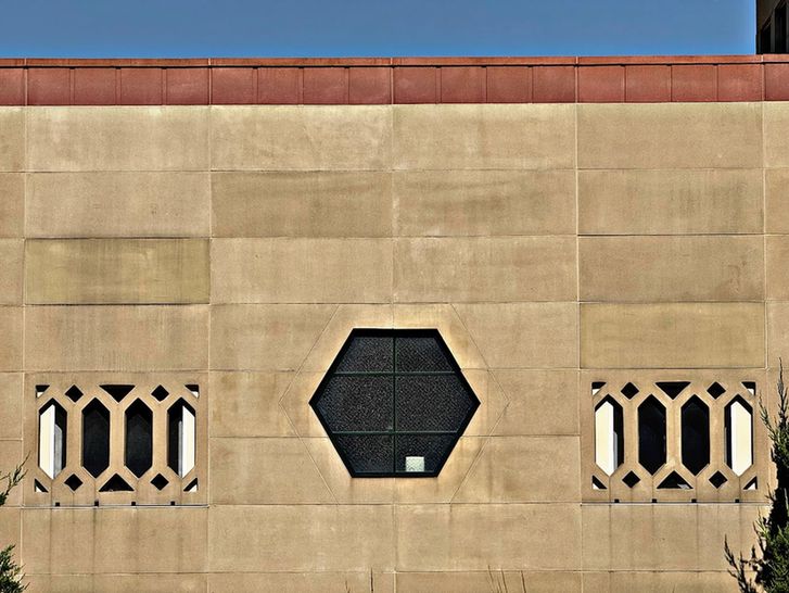Color detail focusing on a centrally located hexagonal multilight window with dark glass set into the tan stone of the facade. This window is flanked on either side by rectangular windows covered by stone panels with decorative grilles.