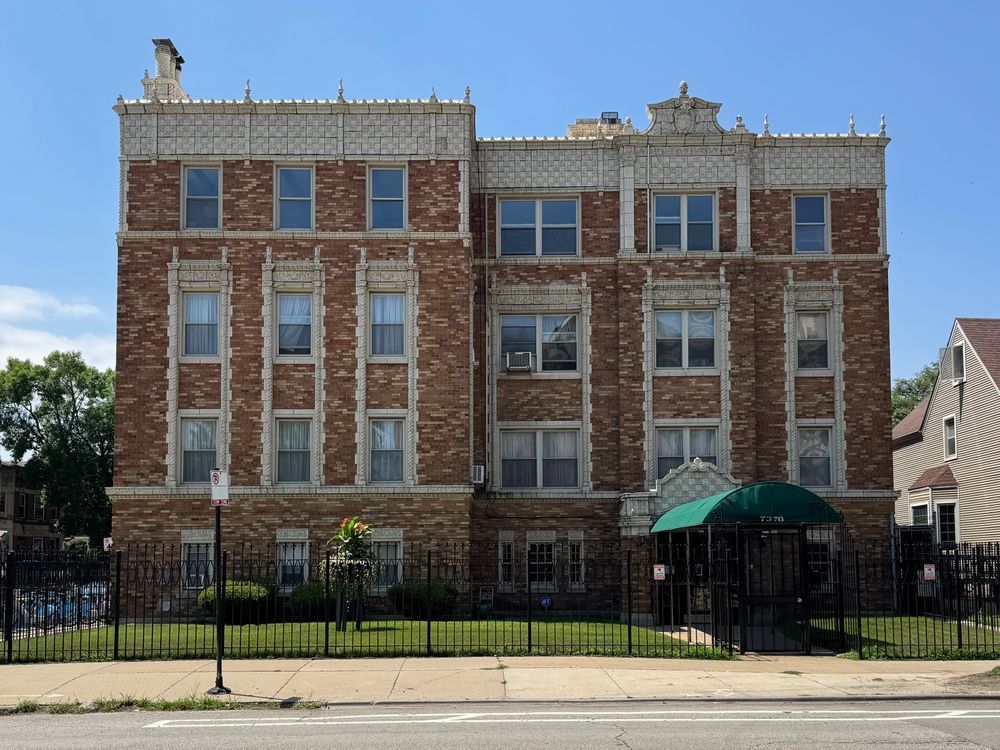 Color photo of a brown variegated brick building with a flat roof and a thicc band of terra cotta up top that is additionally ornamented by finials. Terra cotta also surrounds windows on the first three floors, where decorative lintels abound, and the second the third floors additionally boast two story bands of quoins. 
