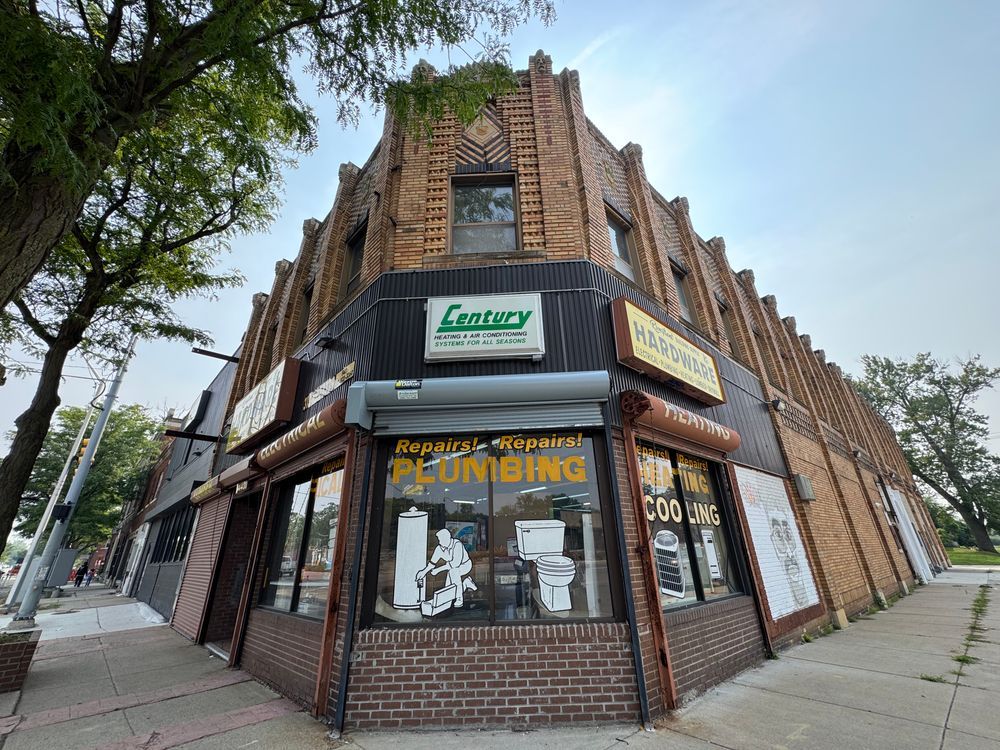 Color photo of a two story building taken from the former corner entrance at a wide angle. It has been partially filled in with nonmatching unsympathetic brick at the bottom, and has large windows with drawings of toilets and guys doing HVAC work, advertising the business currently housed here.
