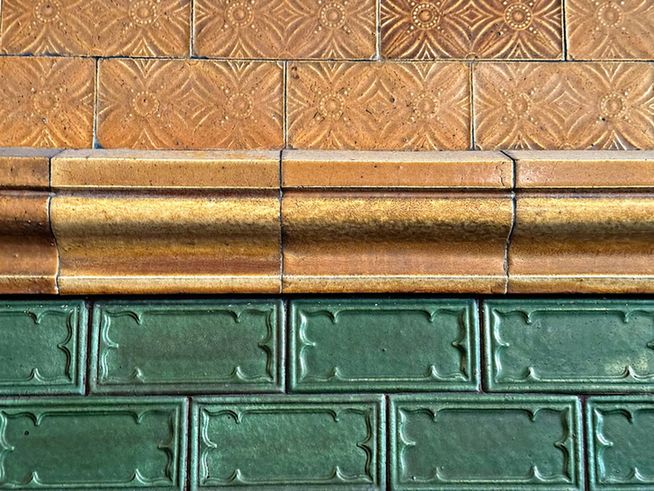 Color detail of a section of wall. At top are highly detailed golden tan tiles embossed with a geometric and floral design. A matching molding separates this from the bottom tiles, which are a gorgeous green and outlined in raised, Gothic tracery.