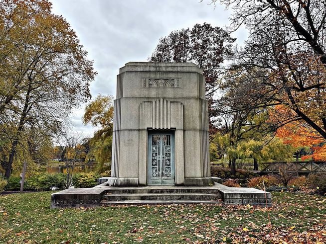 Color photo of a weathered Art Deco mausoleum. Fallen leaves are spread across the grass surrounding it, and in the far background, a pond glows orange, reflecting the changing trees around it. To the right is a wooden bridge leading off to a small island, orange and red bushes and trees surrounding it.