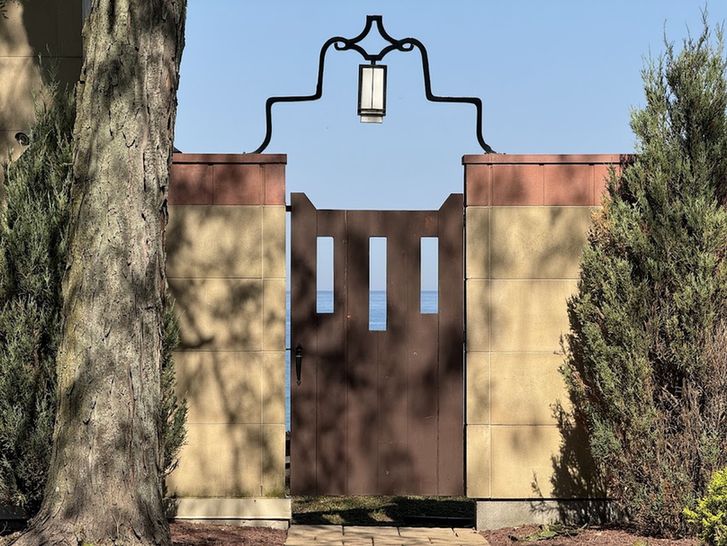 Color close-up of a dark brown wooden gate hung between tan stone walls. The gate has three vertically oriented cut-outs through which can be seen a blue sky and an even bluer Lake Michigan. Above it, a lantern is suspended from black metal scrollwork that arches above the gate.