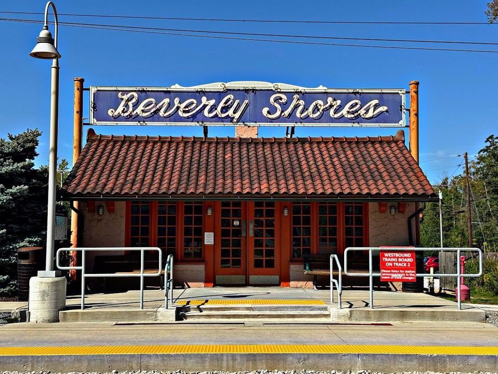 Color photo of the station side of the building taken from across the tracks. Two orange multilight doors are centered between three sets of tall, multilight windows. On top of a red tile roof is an incredible vintage neon sign that has the words Beverly Shores in a white cursive font on a dark blue background.