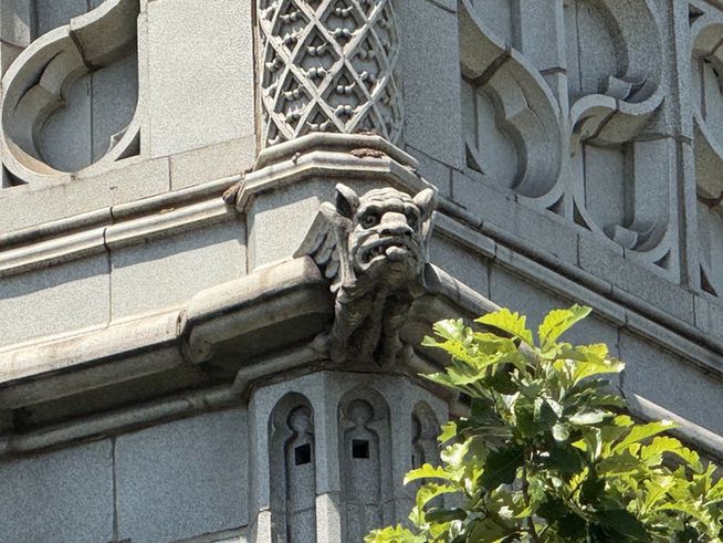 Color close-up of a particularly bilious looking gargoyle precariously extending from a corner of a building and surrounded by all manner of Gothic ornament.
