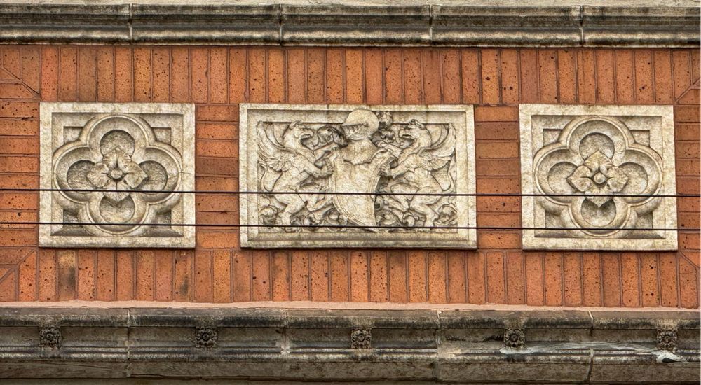 Color close-up of more of the stock ornament on the building. There are 3 panels in total: two quatrefoil panels on either side of a panel featuring a weird knight's helmet on a shield that honestly looks more like a Xenomorph. Completing this panel are two winged heraldic beasts pawing at the helmet/Xenomorph.
