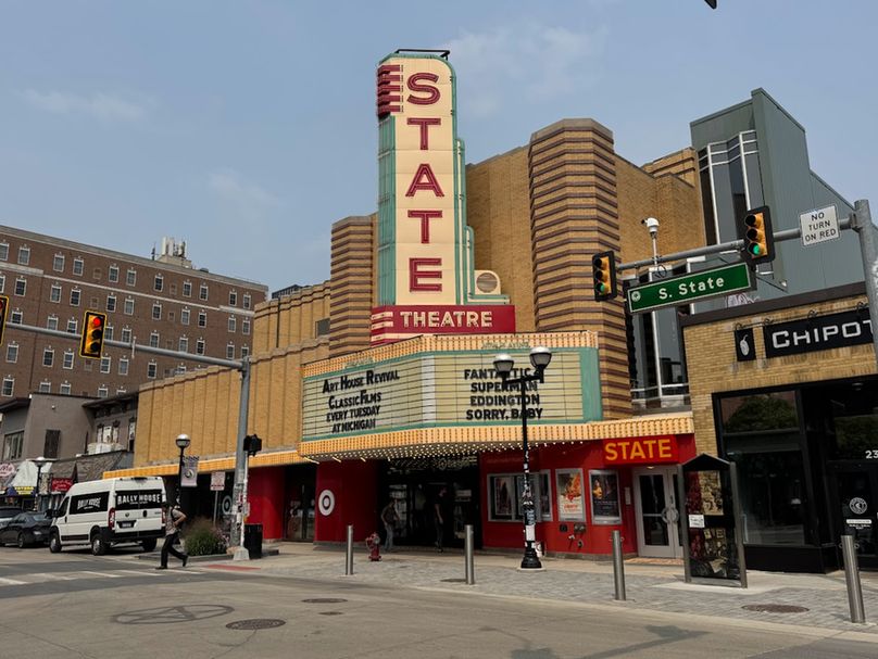 Color photo of an extraordinary theatre that still functions as such, but also has been turned into a Target. I know. It somehow still works. The photo is taken from an angle across the street, showing the marquee and the giant cream, red and light teal vertical sign rises above it proclaiming this the STATE THEATRE. Stretching out to the left is an exquisite, two level folded facade of tan brick. 
