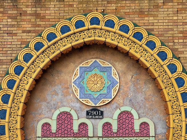 Color close up of the ornament above the entry. Inside of a large horseshoe arch formed by rows of intricately designed polychrome terra cotta fashioned into geometric designs in dark yellow, blue and green, is another incredibly complex circle/star element in a lighter yellow, green and blue terra cotta. Below this, twin designs in pale green and dark pink that seem to mimic in miniature the curving arches common in Islamic architectural designs. Even this element is made extraordinary as it is filled in with tiny, dark pink terra cotta tessellations.