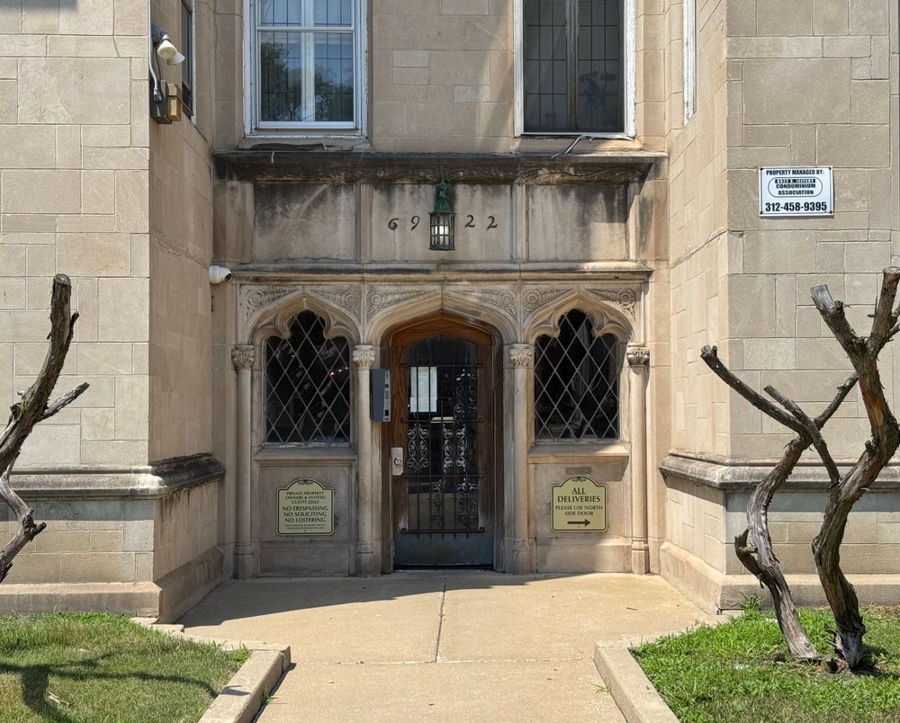 Color photo of the recessed entrance to a light-colored building with a stone base. In the center is a wooden door, flanked on either side by leaded diamond pane windows highlighted by Gothic tracery and above this, lovely foliate stone carvings. Between each of these elements is an engaged column. From either side of the walk leading to the entrance are creepy, naked bushes completely lacking leaves in the middle of summer, taking the Gothic fairy tale thing a little far, if you ask me.