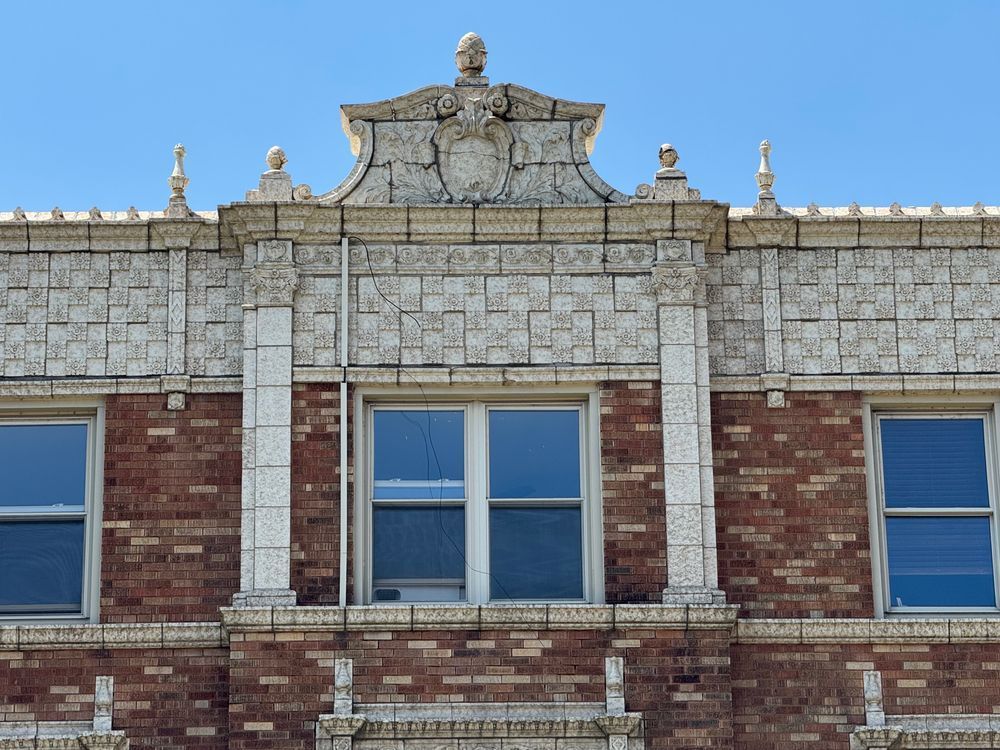 Color close-up of a rather bonkers amount of terra cotta ornamentation at the very top of the building. There are about a million finals, an enormous expanse of small tiles (some plain, and some with a raised rosette) arranged in a checkerboard pattern, pilasters on either side of a pair of windows, and a pediment additionally adorned with a large centered cartouche, plus scrolls and acanthus leaves.