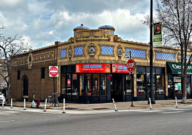 Color photo of part of a row of exquisitely embellished storefronts, taken from across the street. Designed to visually dominate the intersection, it is a riotous combination of decorative elements, all rendered in a gorgeous, warm tan terra cotta with stretches of blue that make it pop all the more.
