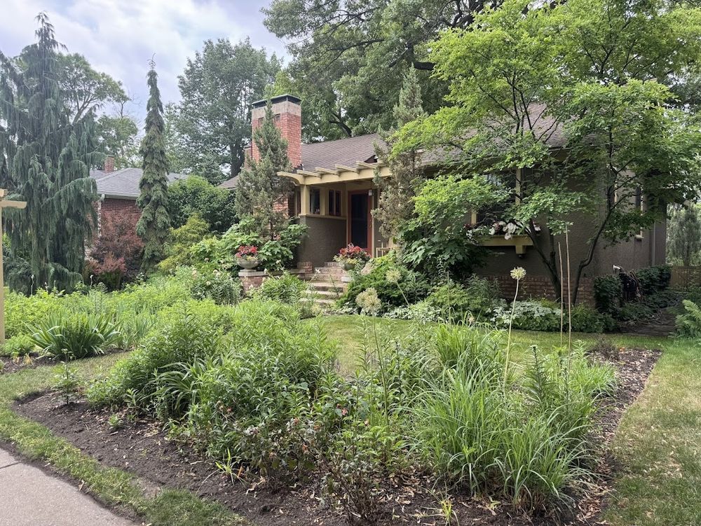 Color photo of a one story home of brown stucco with a brick base, taken from an angle. The front yard is filled with flowers and greenery and the home looks magical.