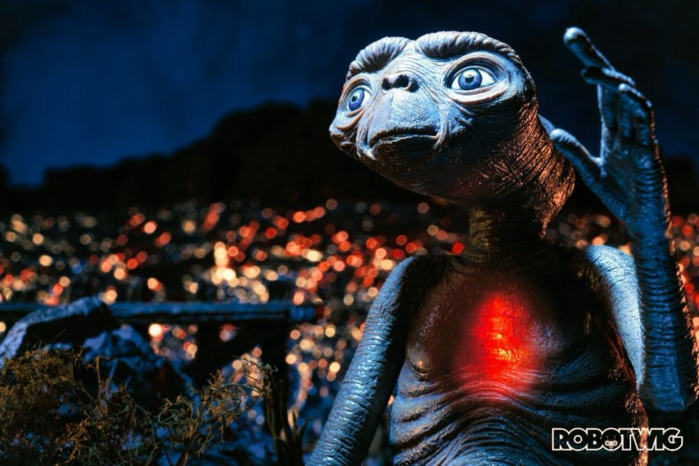 E.T. Looks up at a bright light coming from his ship as it leaves him behind. His chest is glowing red and city lights can be seen in the background 