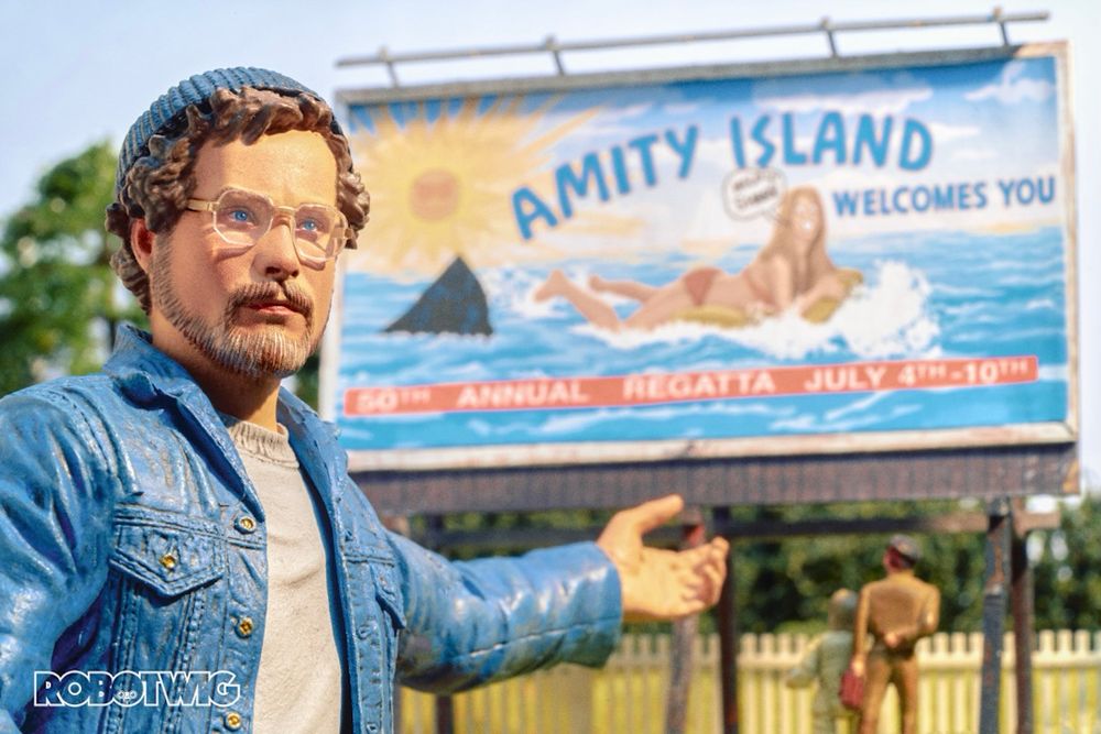 Hooper points toward graffitied billboard with Shark fin painted in it. Billboard says Amity Island Welcomes You 