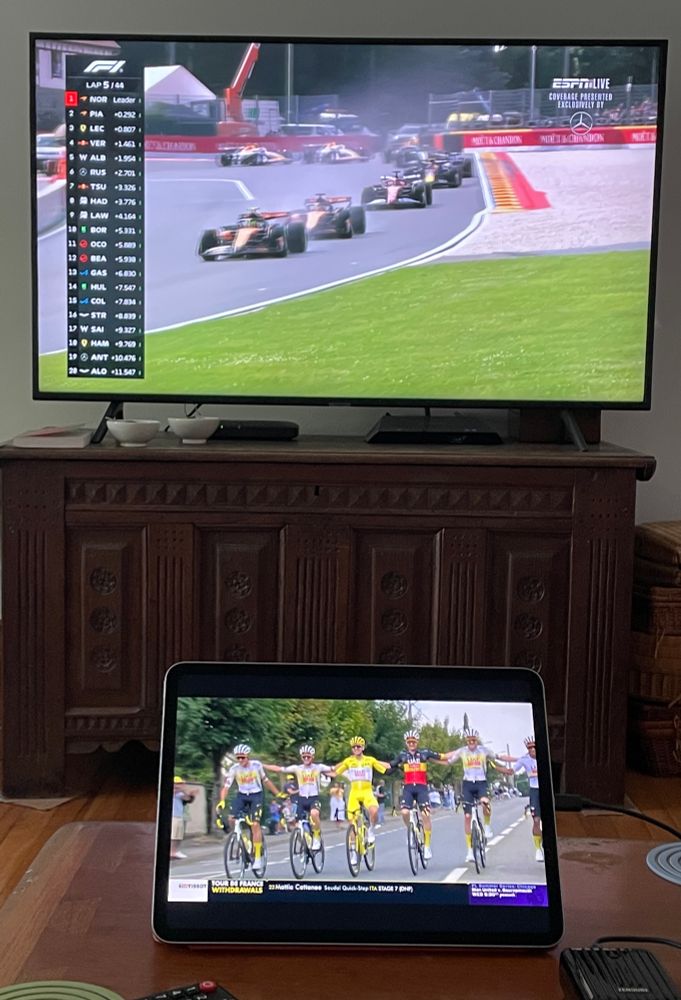 Picture of two screens in a living room. Top TV screen shows first lap of F1 Belgian Grand Prix, bottom screen shows opening kilometers of final stage of 2025 Tour de France with leaders in various classifications  locking arms. 