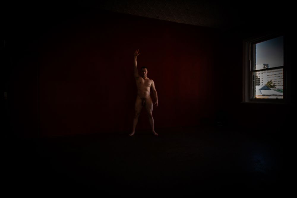 Male model, very dark background, facing viewer with left hand raised in air, centre frame. Window is right of frame