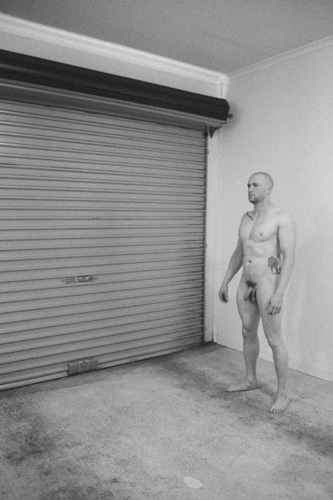 Male nude standing left of frame, facing right of frame, next to a garage door.