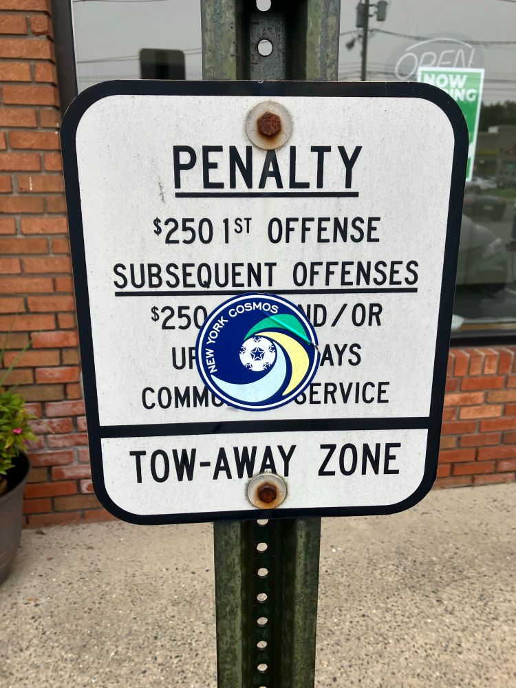 a New York Cosmos sticker over a no parking sign with PENALTY at the top 