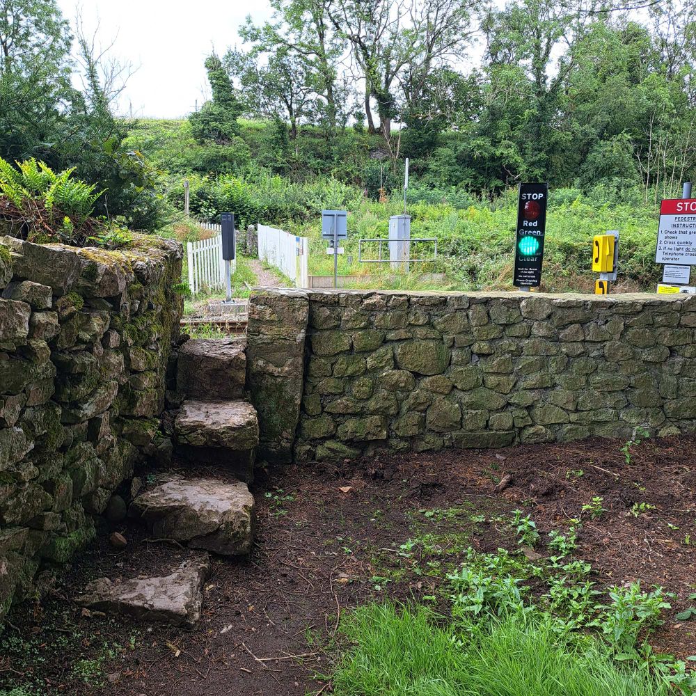 Three large stone steps leading through a wall onto the railway. There is a green traffic light, indicating that it is safe to cross the railway track.
