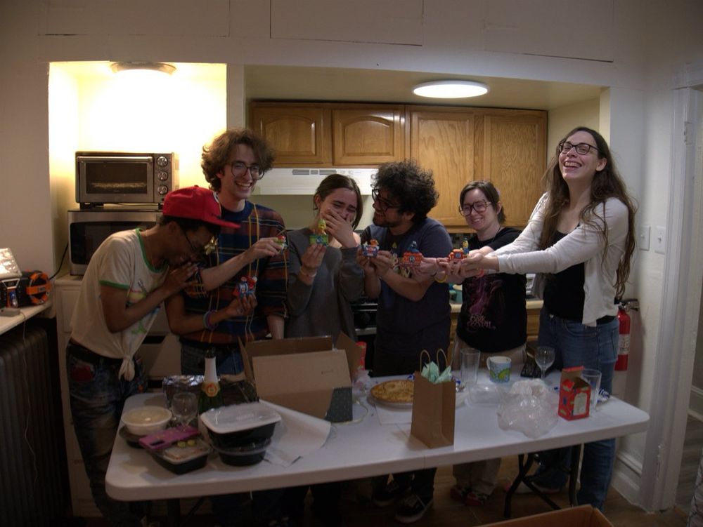 Six people (Nova, Gav, Kate, Richard, Moomin, and Lucy) laughing and holding little Sesame Street decorative train cars around a messy folding table in a small room.