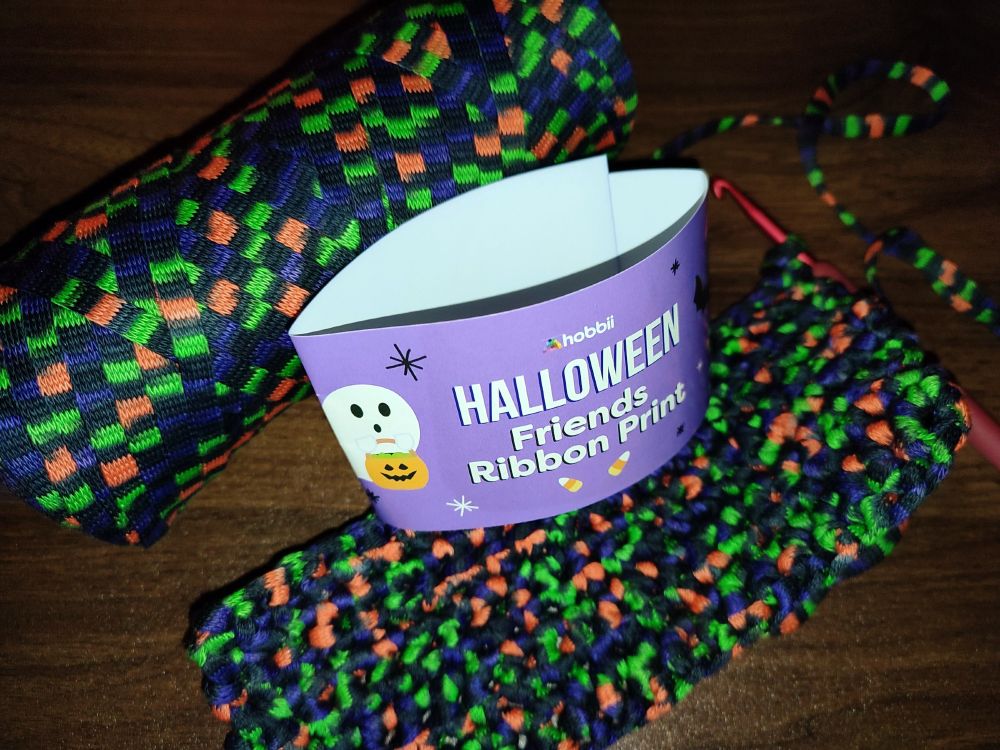 Gehäkeltes Rechteck aus festen Maschen liegt auf einem Tisch. Das verwendete Garn ist schwarz mit orangen, neongrünen und lila Flecken.
Das Knäuel liegt daneben. 
Auf der Banderole steht:
Hobbii
Halloween
Friends Ribbon Print 