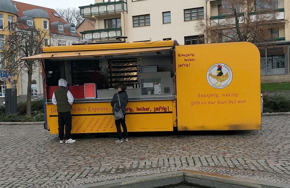 Man sieht einen Verkaufsstand für gegrillte hänchen. Er ist gelb. Zwei Kund*innen stehen davor.